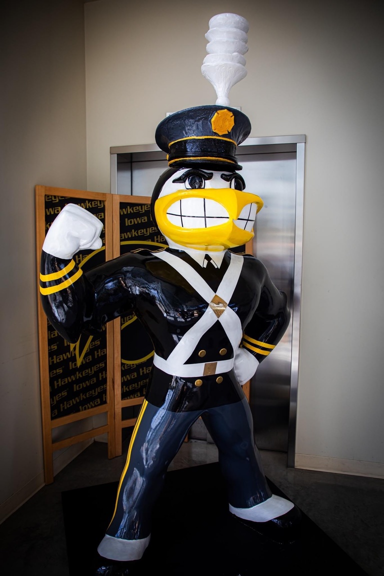Alumni Band Herky fully costumed in the original Marching Band Uniform from the 1940s and 50s. The outfit includes a hat with a tall white feather, a black military looking coat with two gold bands around the wrist, and white straps that cross in the front as well as buckles on Herkys shoulder. He also wears white gloves. 