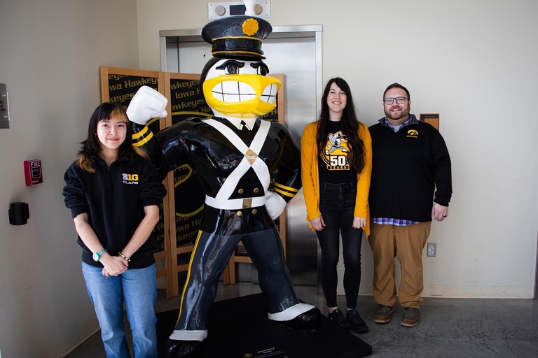 Artists Mae Dunning in black pull-over and jeans, Amanda Stout to Herky's right in yellow cardigan over the Alumni Band's 50th Anniversary t-shirt, and Marching Band Director Dr. Eric Bush in black pull over and khaki pants posing with the completed Alumni Band Herky for Herky on Parade. The Alumni Marching Herky is center, fully costumed in the original Marching Band Uniform from the 1940s and 50s. 