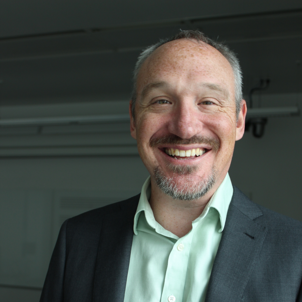 Dr. Joshua Albrecht in a Voxman Music Building classroom, smiling at the camera in a green shirt and gray blazer