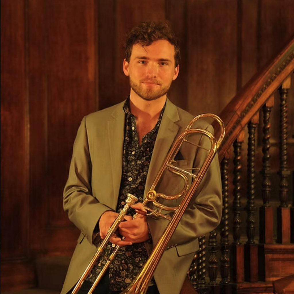 Noah Perkins, pictured holding his trombone with a wood paneled wall and staircase behind him