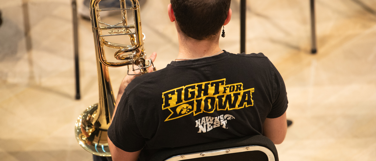 a student brass musician shown from behind, in a black t-shirt that say's "fight for iowa" in gold letters