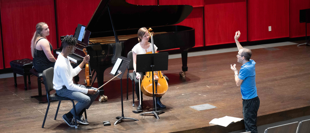 students playing piano, violin, and cello, on stage working with a professor 