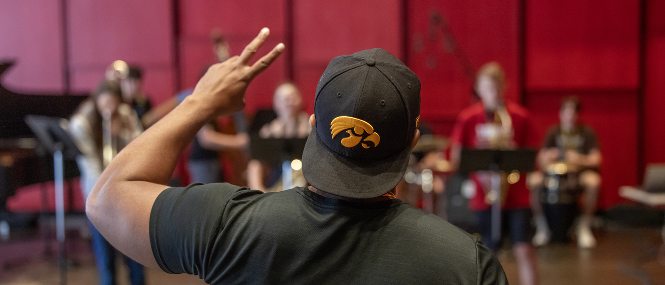 professor william menefield, viewed from behind wearing a hawkeye baseball cap turned backward. He's holding up two fingers to the students in front of him holding their instruments