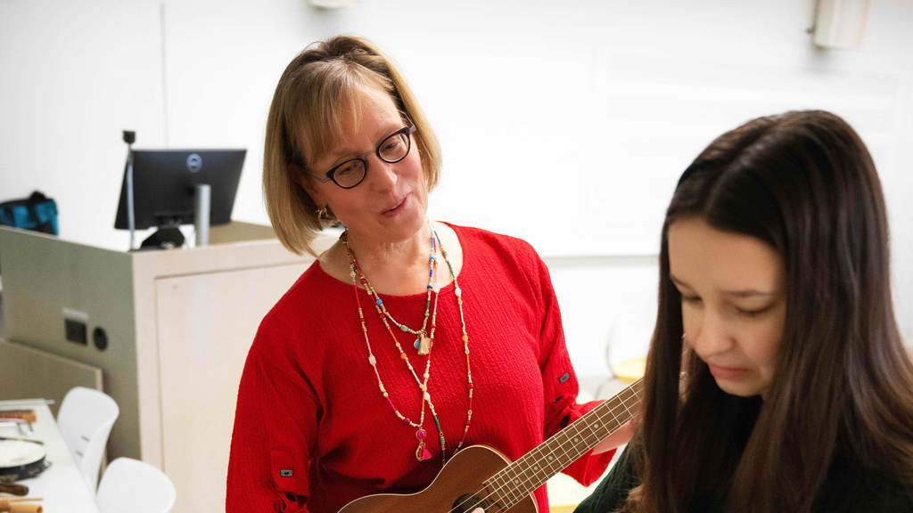Music Therapy blonde woman professor wearing a bright red sweater and holding a guitar, talking with a female student also holding a guitar