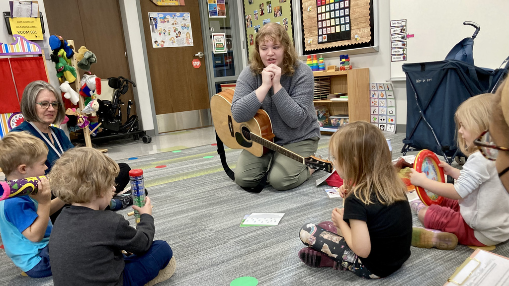 A female music therapist sitting with a guitar on the floor of a classroom, surrounded by small children