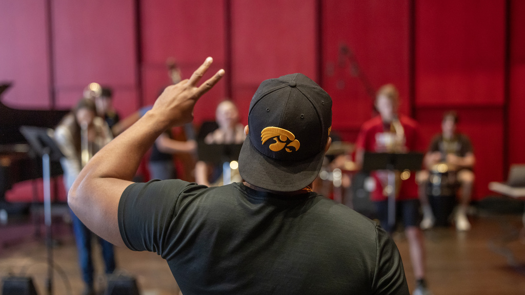 professor william menefield, viewed from behind wearing a hawkeye baseball cap turned backward. He's holding up two fingers to the students in front of him holding their instruments